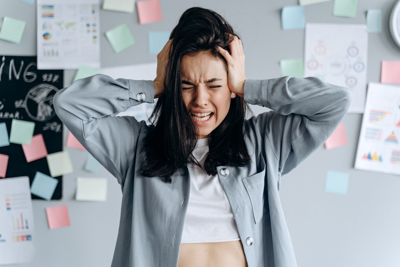 A woman in a grey shirt looking stressed in front of a wall of charts and sticky notes.